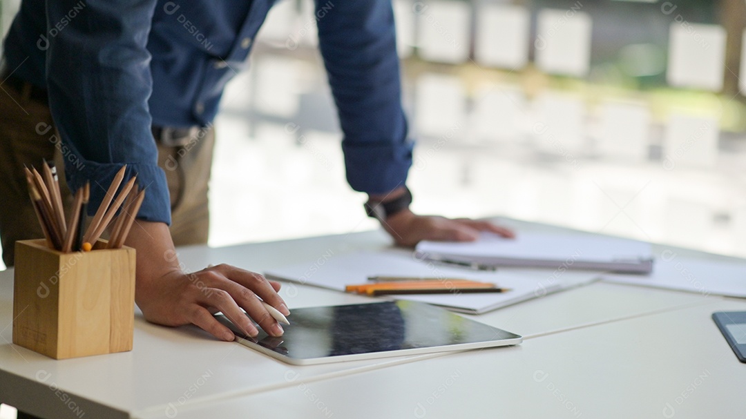 Foto recortada de um jovem segurando uma caneta para usar um tablet na mesa com artigos de papelaria em um escritório contemporâneo.