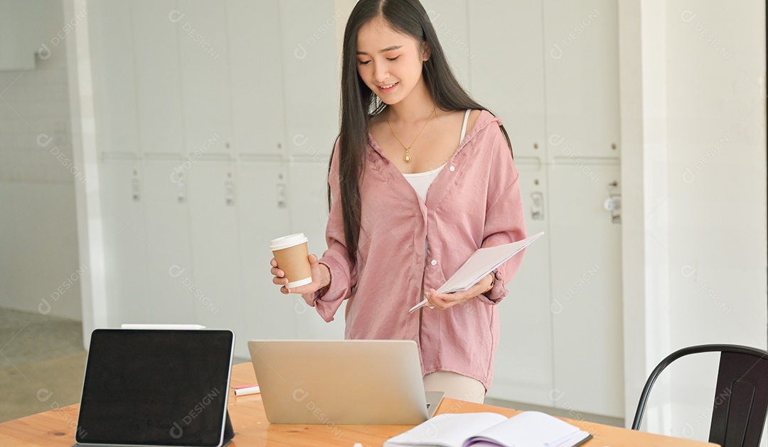 Aluna segurando uma xícara de café e documento de mão com um laptop em cima da mesa. Ela está acelerando sua tese para se formar.