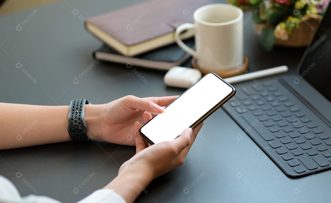 Jovem usando um smartphone de tela em branco com um laptop e café na mesa.