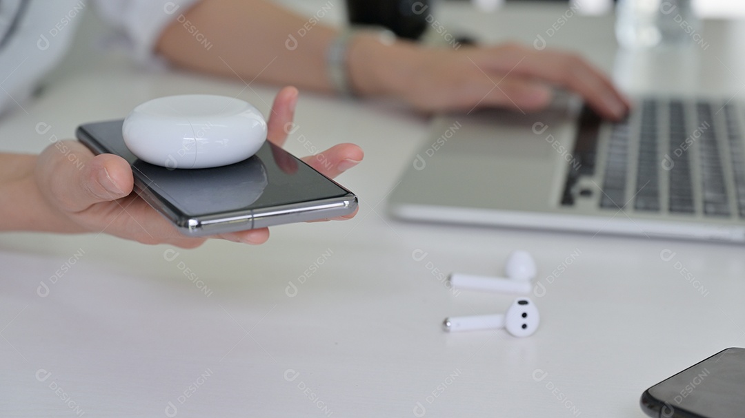 Mulher jovem colocando fones de ouvido em um smartphone para compartilhar um carregador sem fio enquanto ela está usando um laptop.