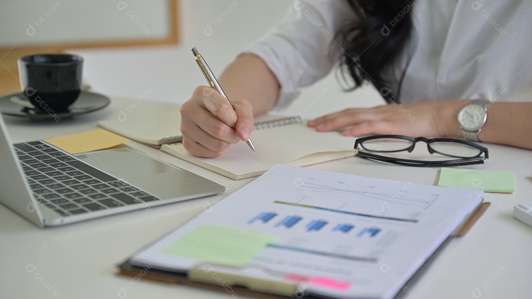 Menina com uma caneta na mão está gravando o desempenho da empresa com gráfico e laptop na mesa.