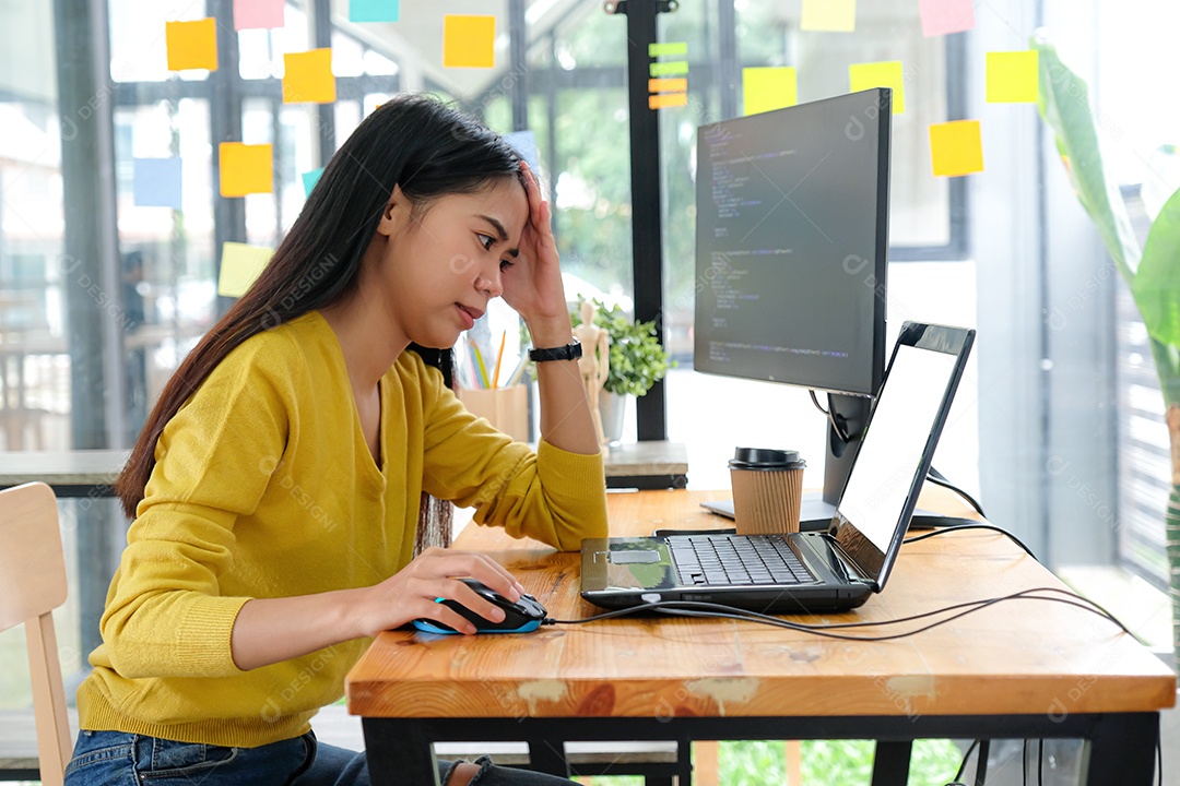 Programadora asiática veste uma camisa amarela, olha para a tela do laptop e mostra uma pose séria.