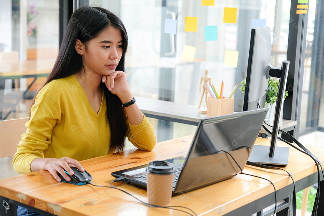 Programadora asiática em camisas amarelas estão usando laptops e PCs. Ela colocou as mãos no queixo e ficou séria.