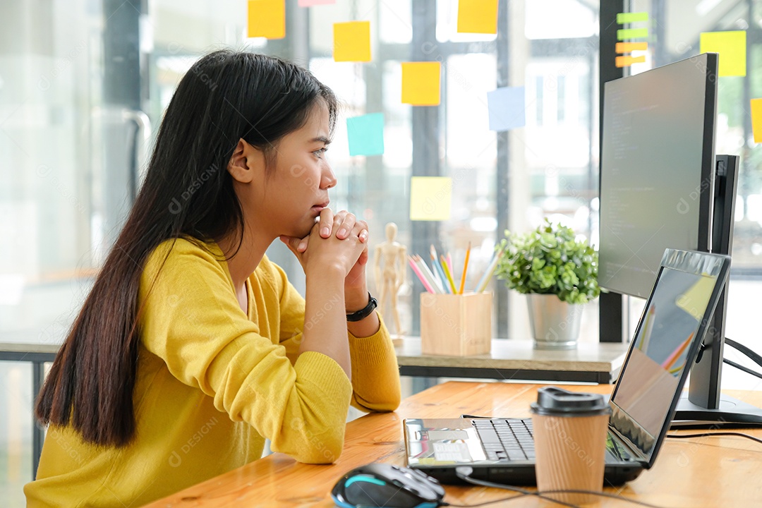 Programadora asiática para camisa amarela sentada com a mão no queixo, ela olhou para a tela do computador e ponderou.