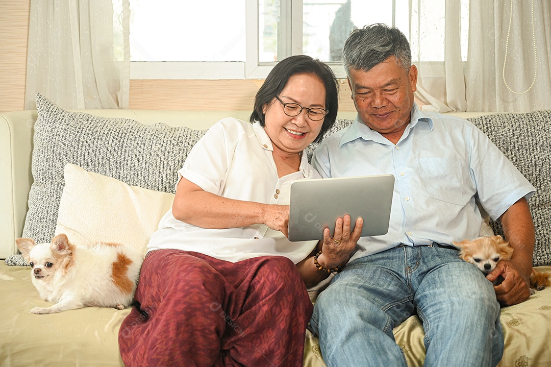 Mulher idosa e um homem asiático sentados em um sofá estão usando um tablet. Eles sorriram alegremente, o cachorro Chihuahua sentado ao lado.