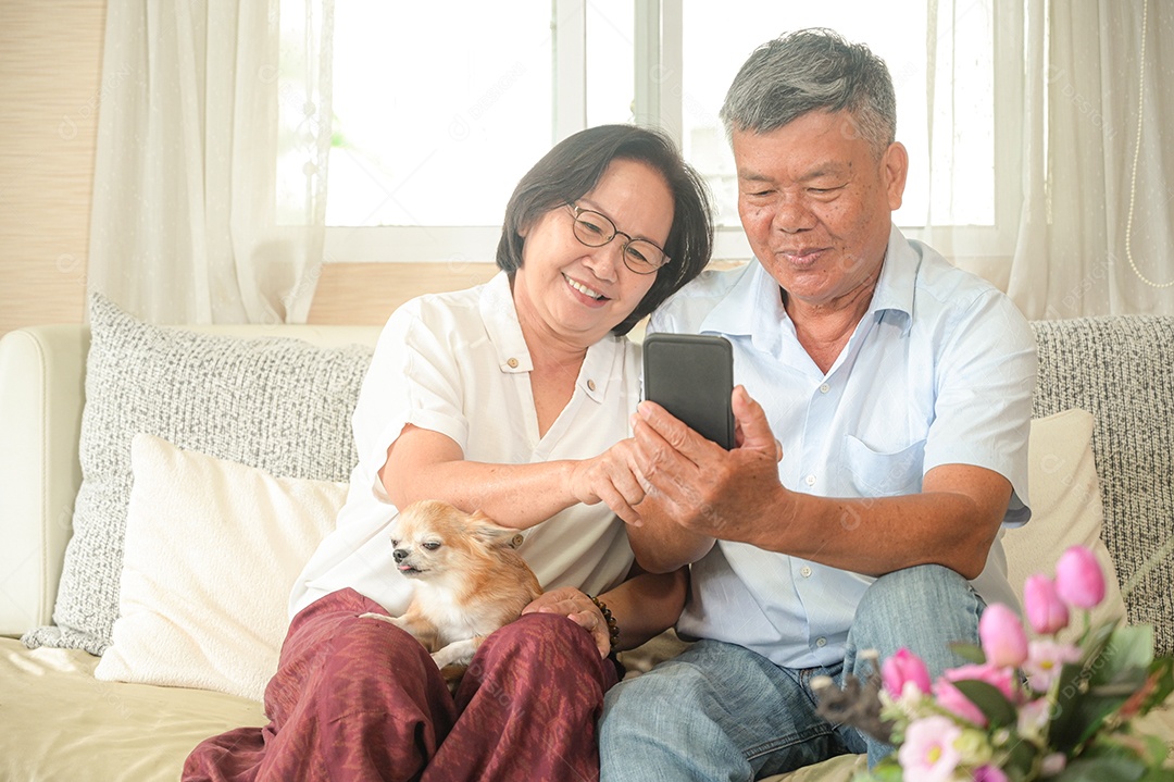 Mulher e homem asiáticos estão sentados no sofá. Eles estão segurando um smartphone. Eles estão sorrindo com uma chamada de vídeo.