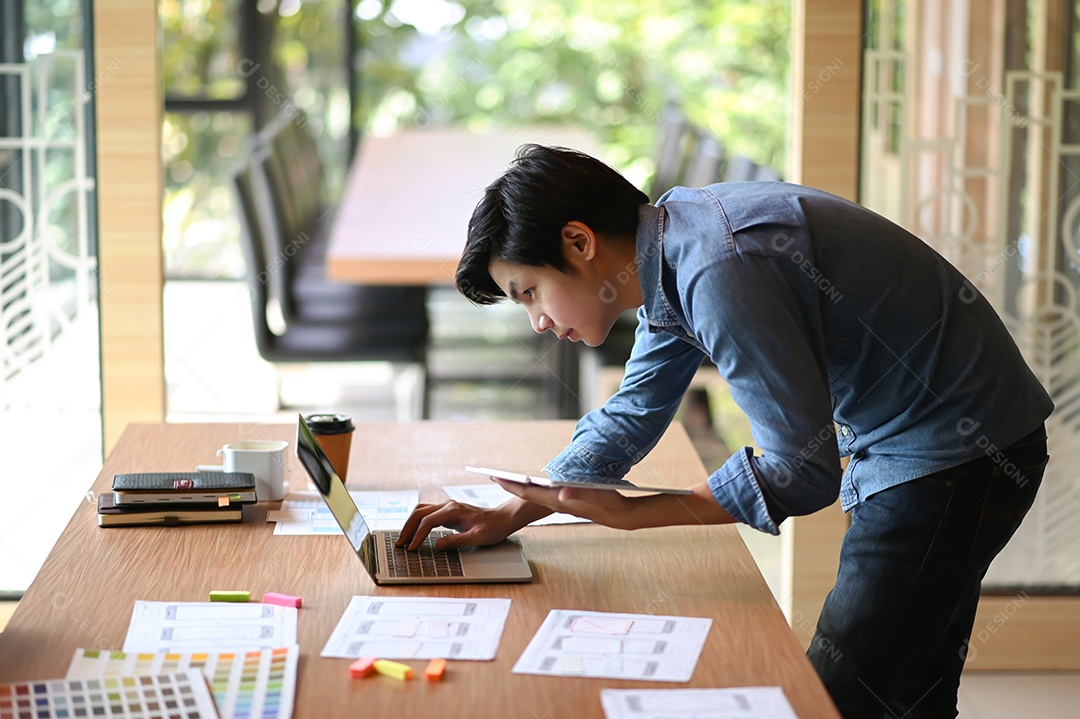 Young designer working in an office. He leaned over to use the laptop and held the tablet in his hand.