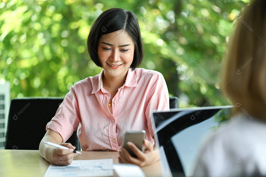 Nova empresária sentada no escritório usando um telefone celular e sorrindo, colegas estão usando laptop sentado em frente.
