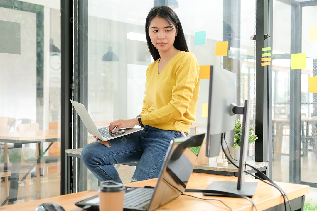 Programador feminino está usando laptop para colocar na perna e criar programas para clientes. Ela sentou na prateleira e olhou para a tela do computador em sua mesa.