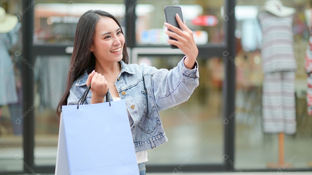 Compradora asiática está usando um smartphone para tirar uma selfie. Ela se levantou e segurou uma sacola de compras.