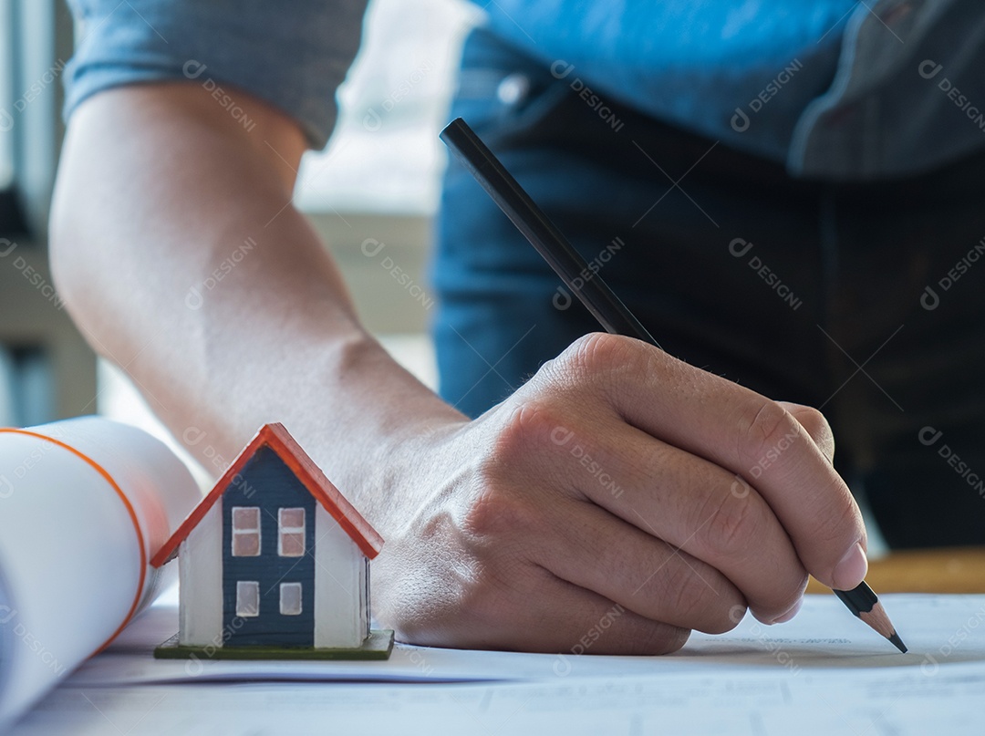 Jovem arquiteto usando um lápis preto para delinear a planta da casa. Casas modelo e plantas de casas são colocadas ao lado.