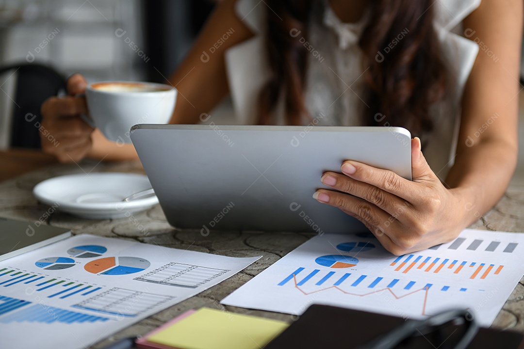 Executivo feminino bebe café e usa tablet durante o intervalo. Ela sentou-se à janela no escritório. Gráfico de dados colocado sobre a mesa.