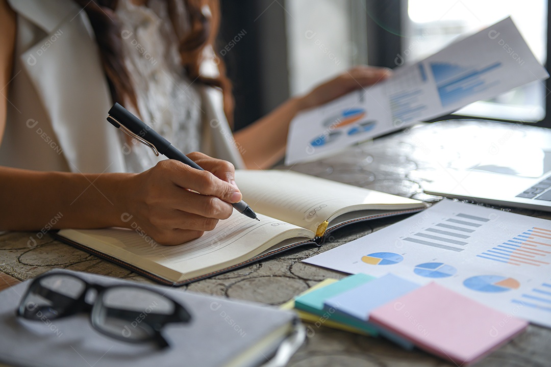 Mulher executiva estão verificando dados de gráficos e tomando notas. Ela segura caneta e gráfico. documentos, cadernos e óculos na mesa.