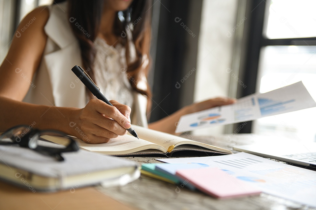 Executivos estão verificando dados de gráficos e tomando notas. Ela segura um gráfico na mão e escreve uma caneta no caderno.