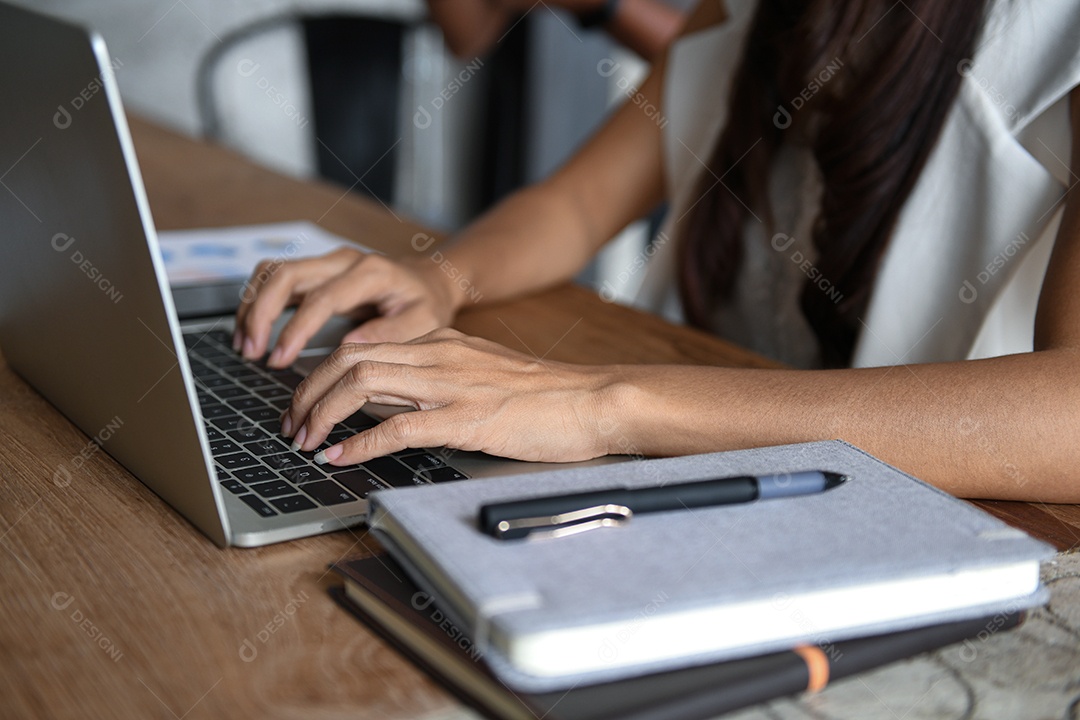 Executivos estão usando laptop. Ela colocou a mão no teclado. A frente tem um caderno e uma caneta colocados sobre a mesa.