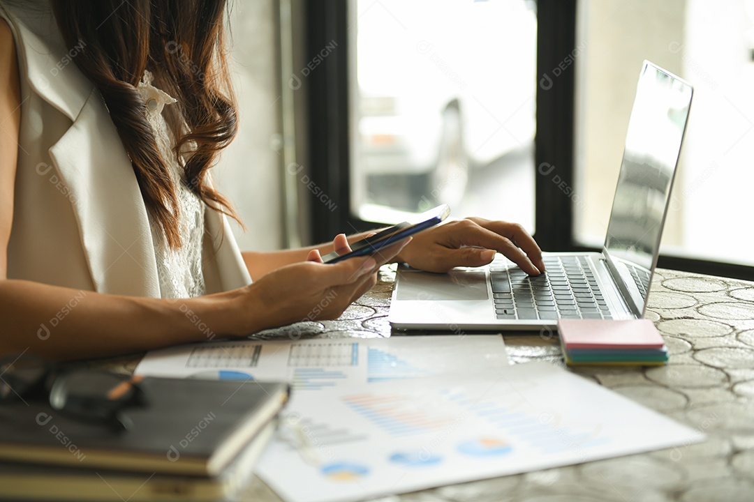 Mulher de negócios estão verificando o gráfico de desempenho da empresa. Ela segura um smartphone e usa um laptop no escritório.