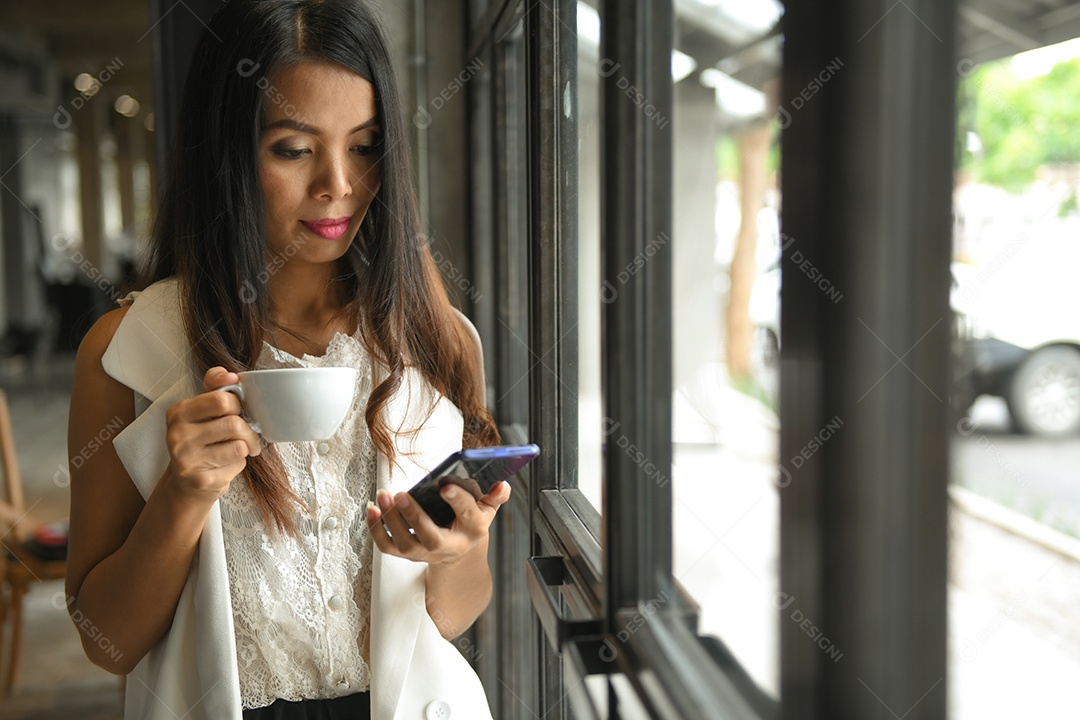 Mulher asiática em um vestido branco segurando uma xícara de café na mão está usando um telefone celular.