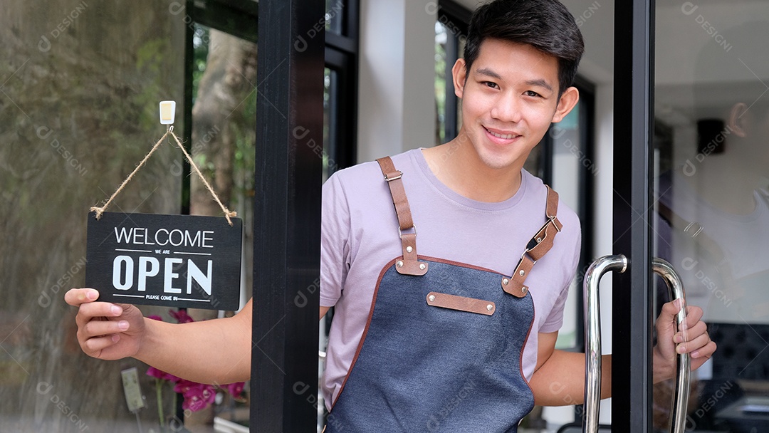 Jovem barista asiático abre uma porta de vidro para receber os clientes. Ele sorri para receber os clientes de maneira amigável.