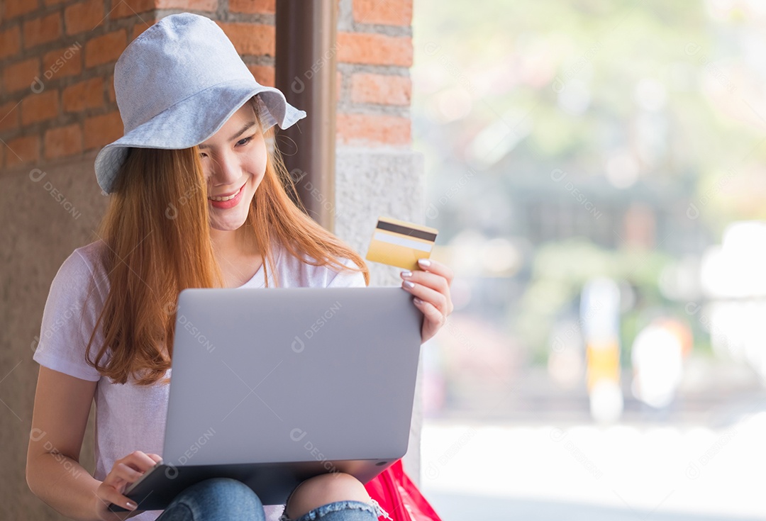 Jovem mulher usando laptop com cartão de crédito na mão para fazer compras on-line, Mulher usa chapéu e sentada no chão, Laptop colocado nas pernas, Conceitos de compras on-line.