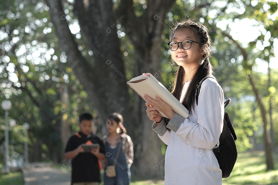 Aluna segura um livro e fica sorrindo no jardim da escola. Ela veste uma camisa branca e usa óculos.