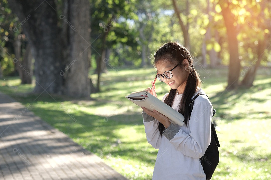 Aluna segura um livro e fica sorrindo no jardim da escola. Ela veste uma camisa branca e usa óculos.