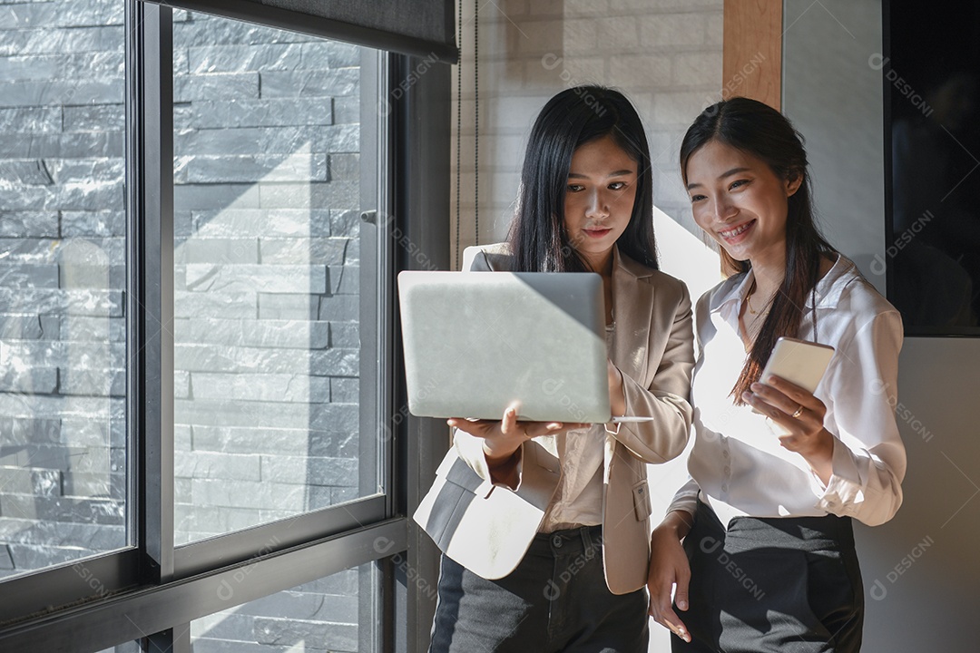 Duas funcionárias estão usando laptop. Ela fica na janela.