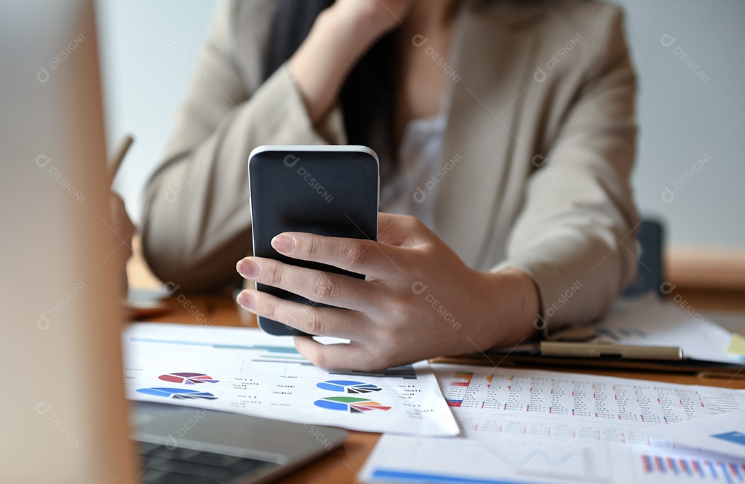 Funcionários estão usando smartphone e laptop na frente, caneta e documentos na mesa.