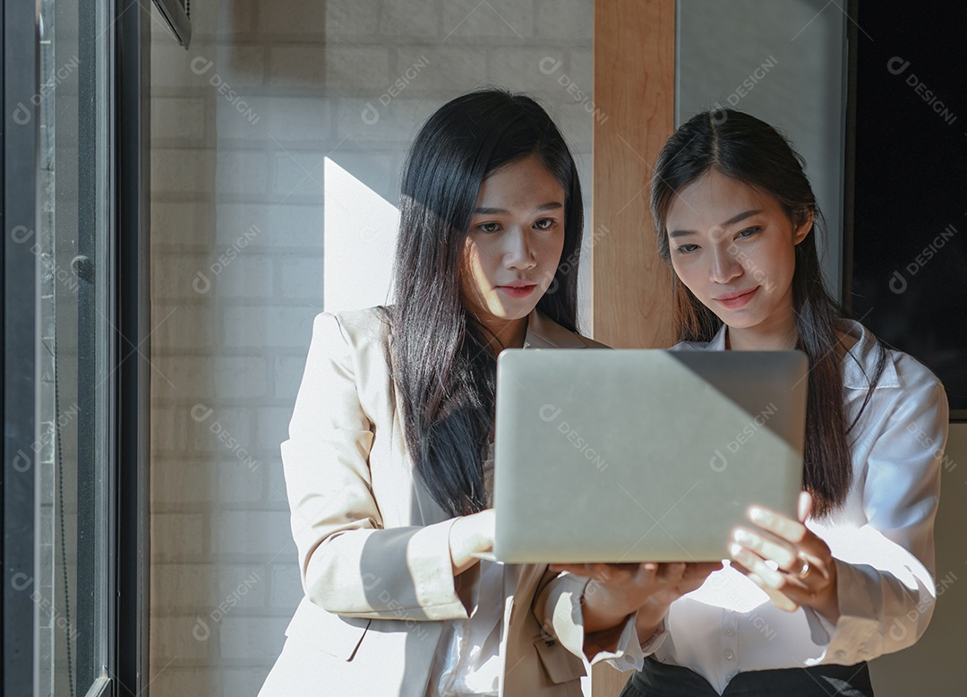 Duas funcionárias estão usando laptop. Ela fica na janela.