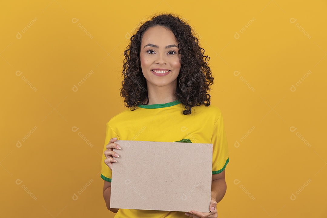 Mulher jovem usando camiseta da seleção brasileira futebol cabelo cacheado sobre fundo amarelo