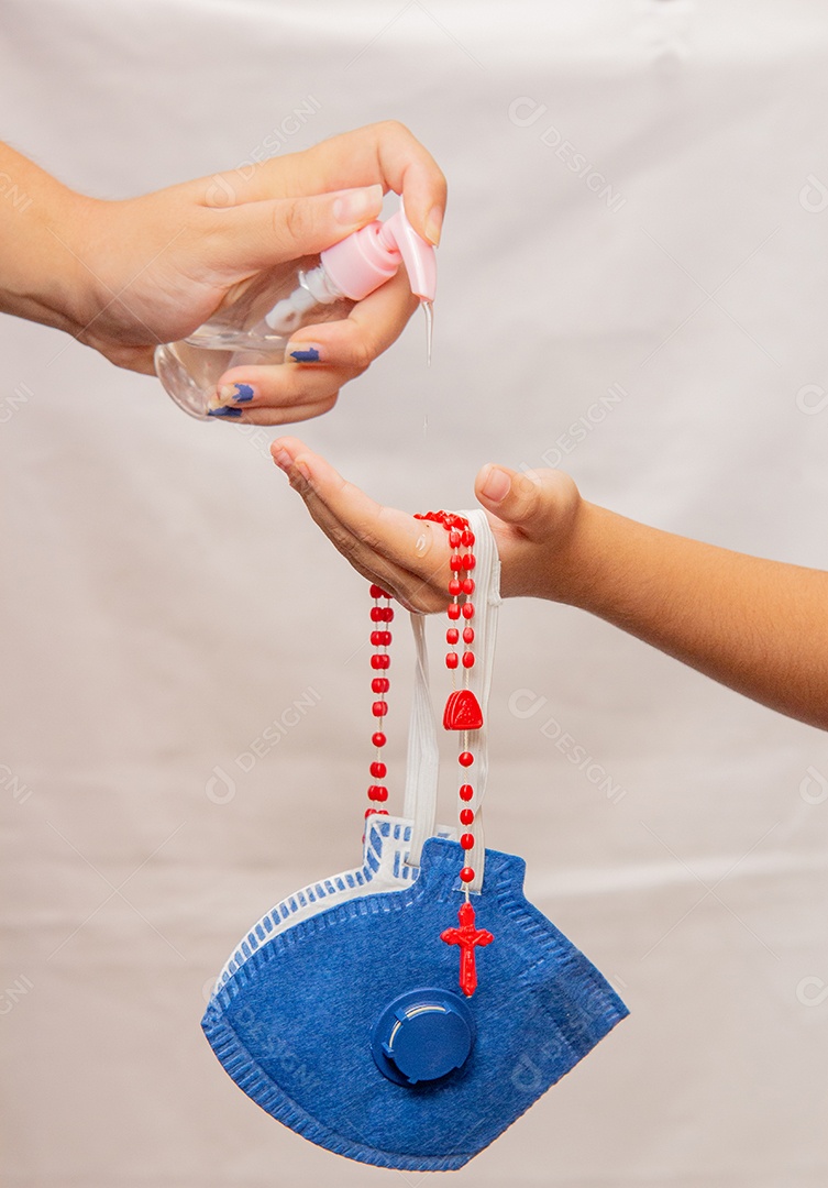 Mulher colocando álcool em gel nas mãos de criança, com máscara e terço vermelho para proteção contra coronavírus no Brasil
