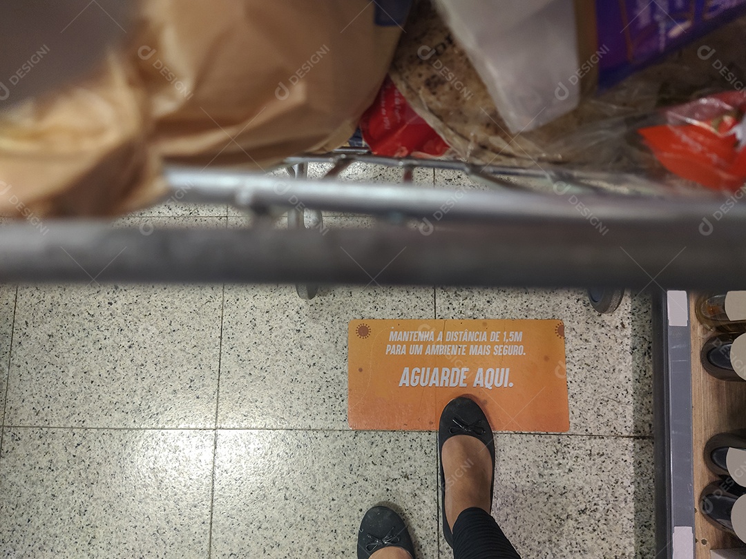 pés femininos ao lado de um carrinho de compras com uma placa no chão escrita em português (mantenha a distância de 1,5 metros