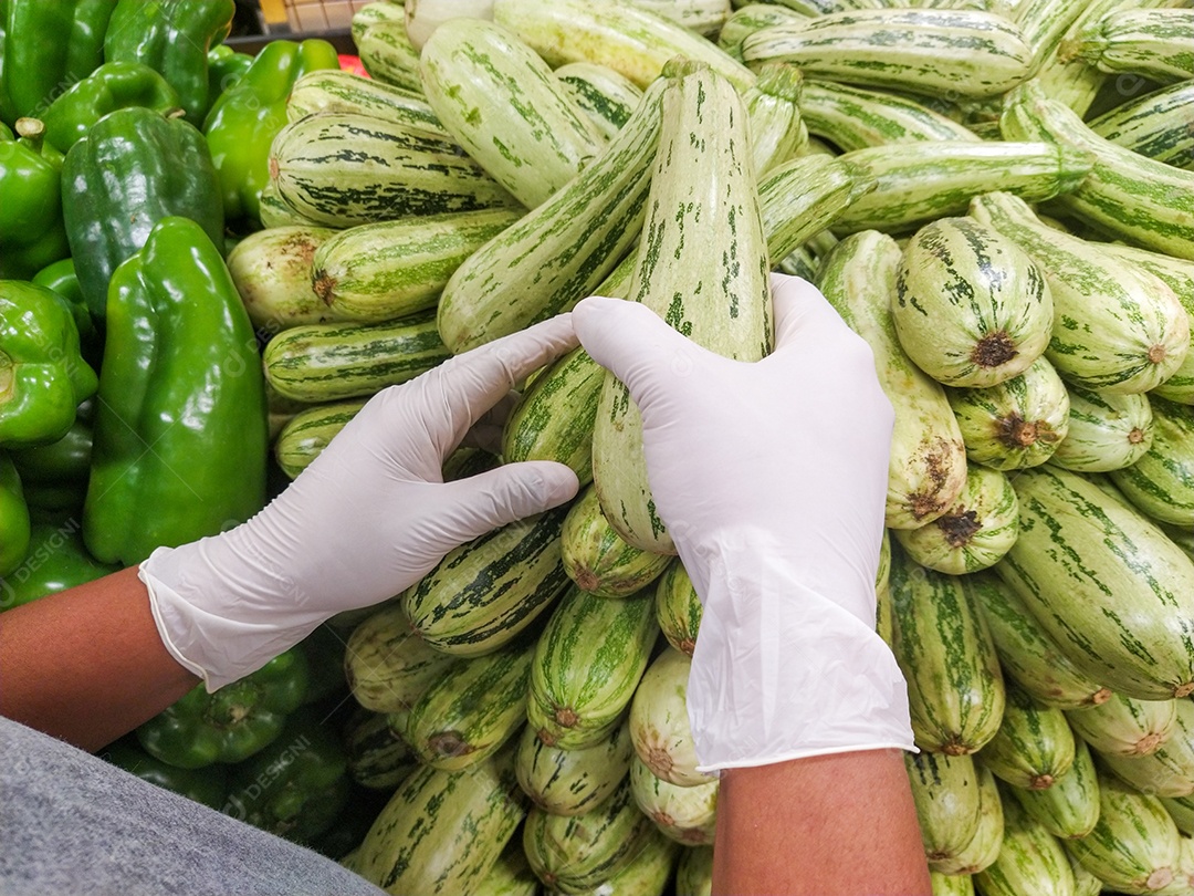 mãos negras de um homem escolhendo abobrinhas com luvas, proteção contra coronavírus no Brasil.