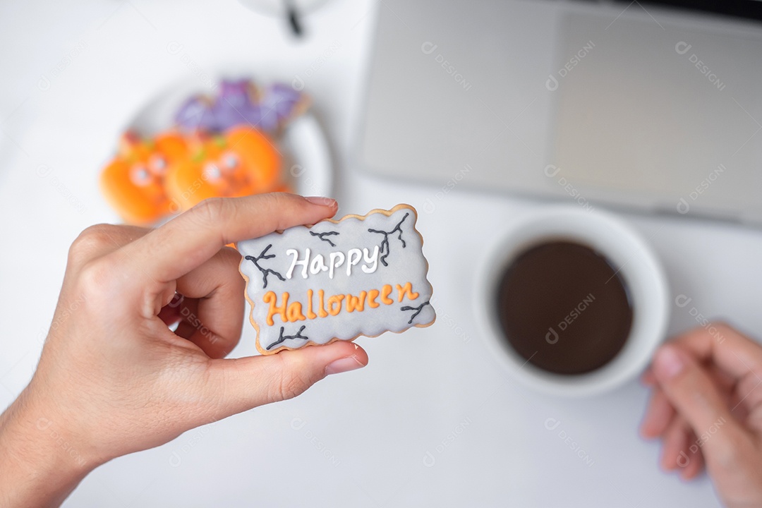 Mão segurando o biscoito de Halloween e tomando café durante o uso do computador portátil. Feliz dia das bruxas, compras online, Olá outubro, outono outono, festivo, festa e conceito de férias