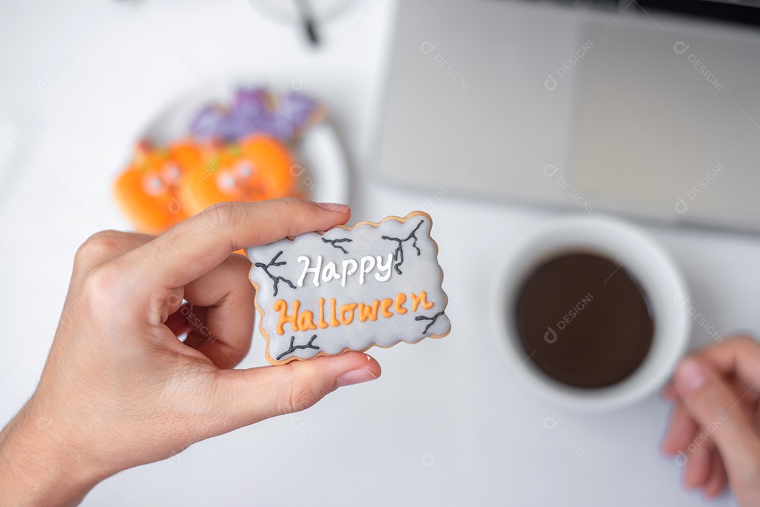 Mão segurando o biscoito de Halloween e tomando café durante o uso do computador portátil. Feliz dia das bruxas, compras online, Olá outubro, outono outono, festivo, festa e conceito de férias