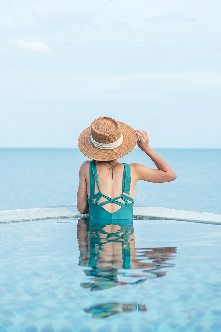 Mulher feliz em traje de banho verde e chapéu na piscina de luxo.
