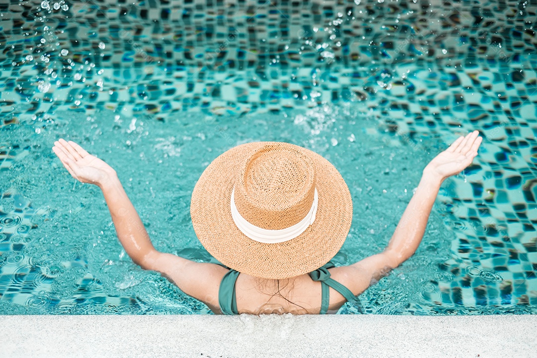 Mulher feliz em traje de banho verde e chapéu na piscina de luxo.