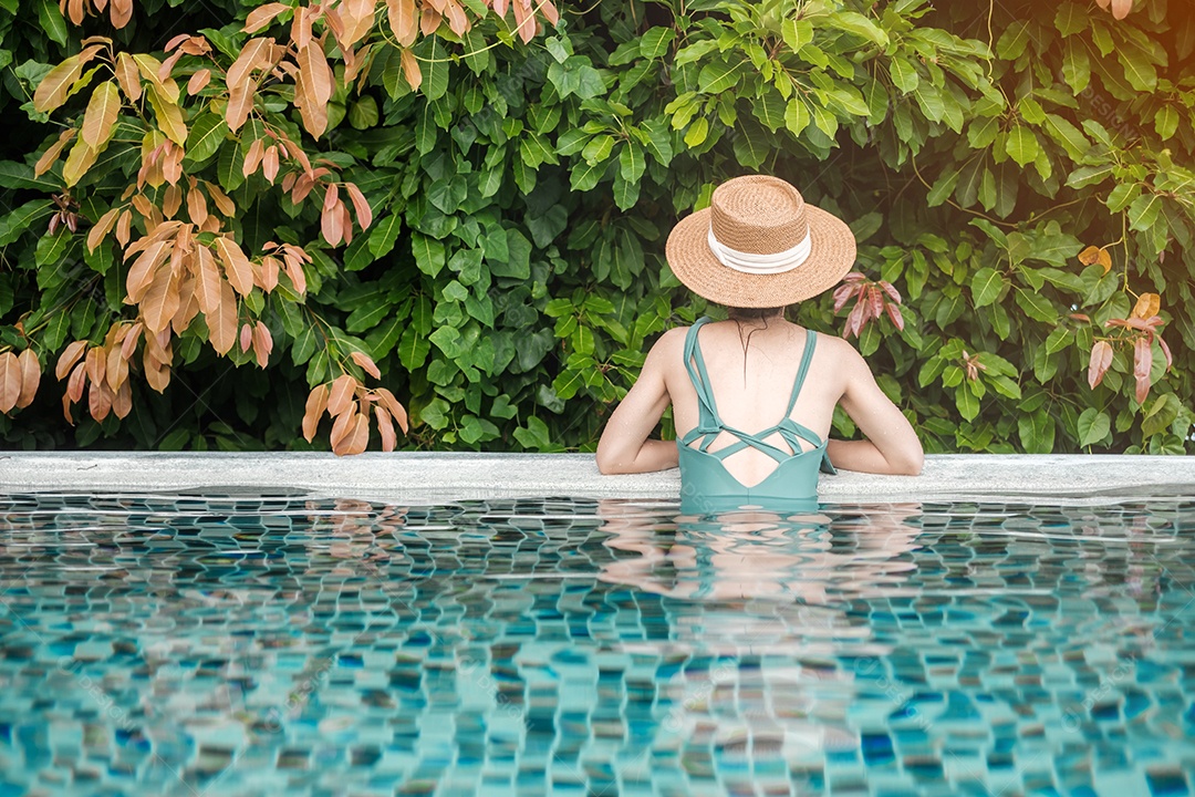 Mulher feliz em traje de banho verde e chapéu na piscina de luxo.