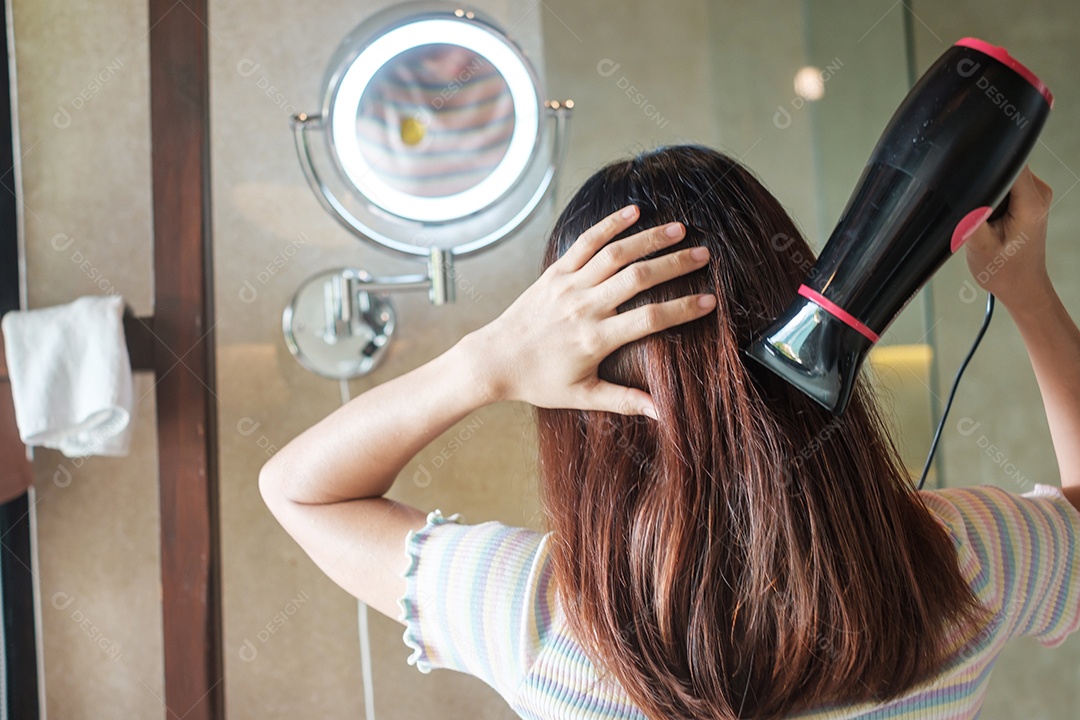 Jovem mulher usando secador de cabelo perto do espelho em casa ou hotel.