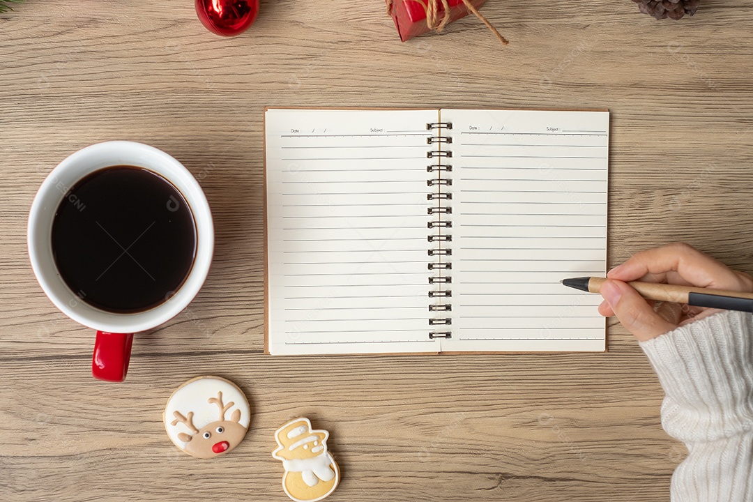 Mão de mulher escrevendo no notebook com xícara de café preto e Natal.