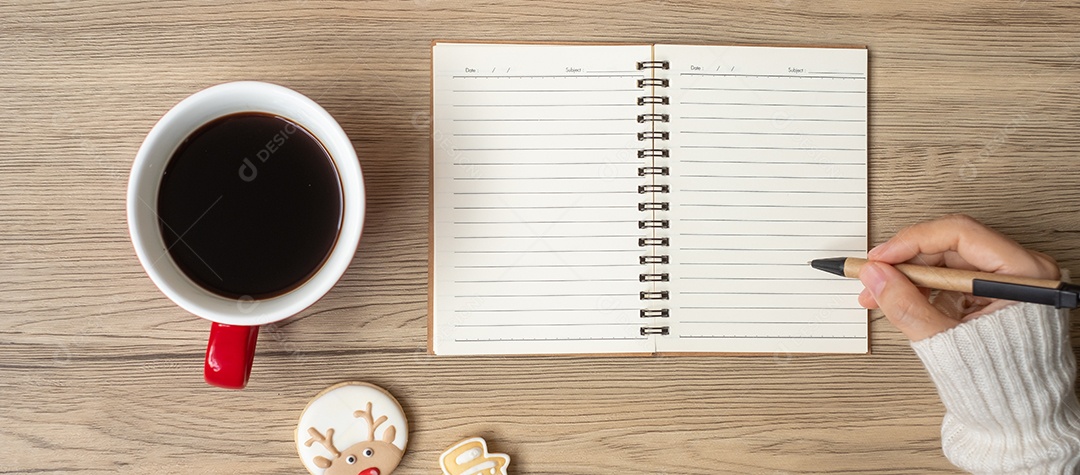 Mão de mulher escrevendo no notebook com xícara de café preto e Natal.