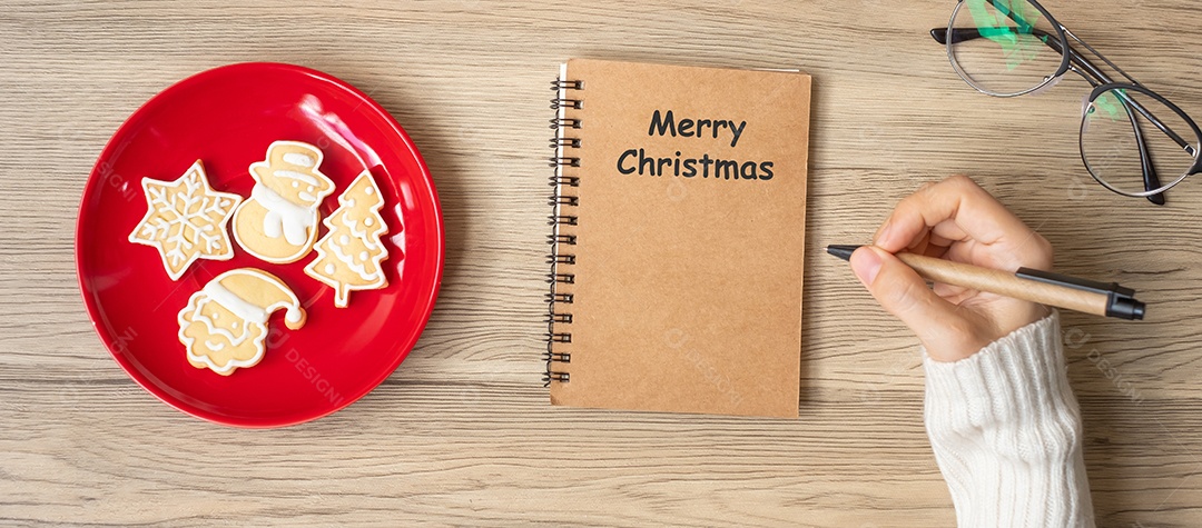 Mão de uma mulher escrevendo no notebook com biscoitos de Natal na mesa.