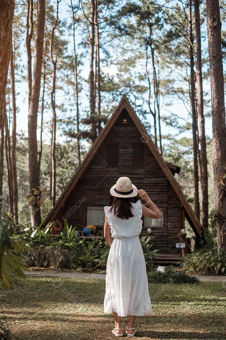 Mulher feliz de vestido branco e chapéu viajando na floresta de pinheiros.