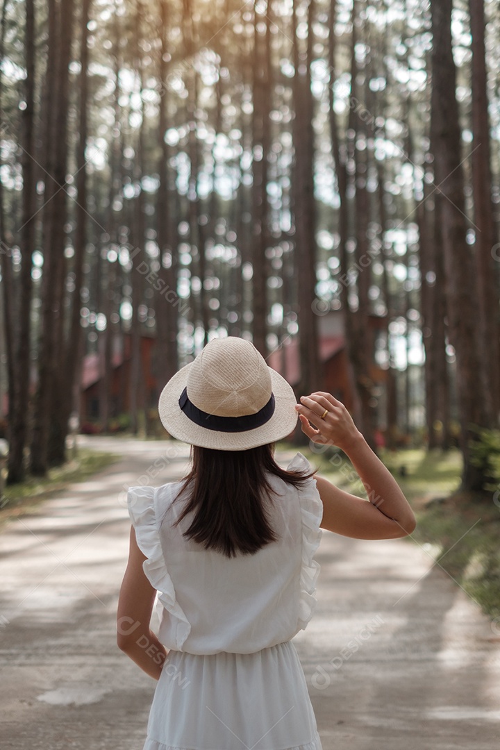 Mulher feliz de vestido branco e chapéu viajando na floresta de pinheiros.