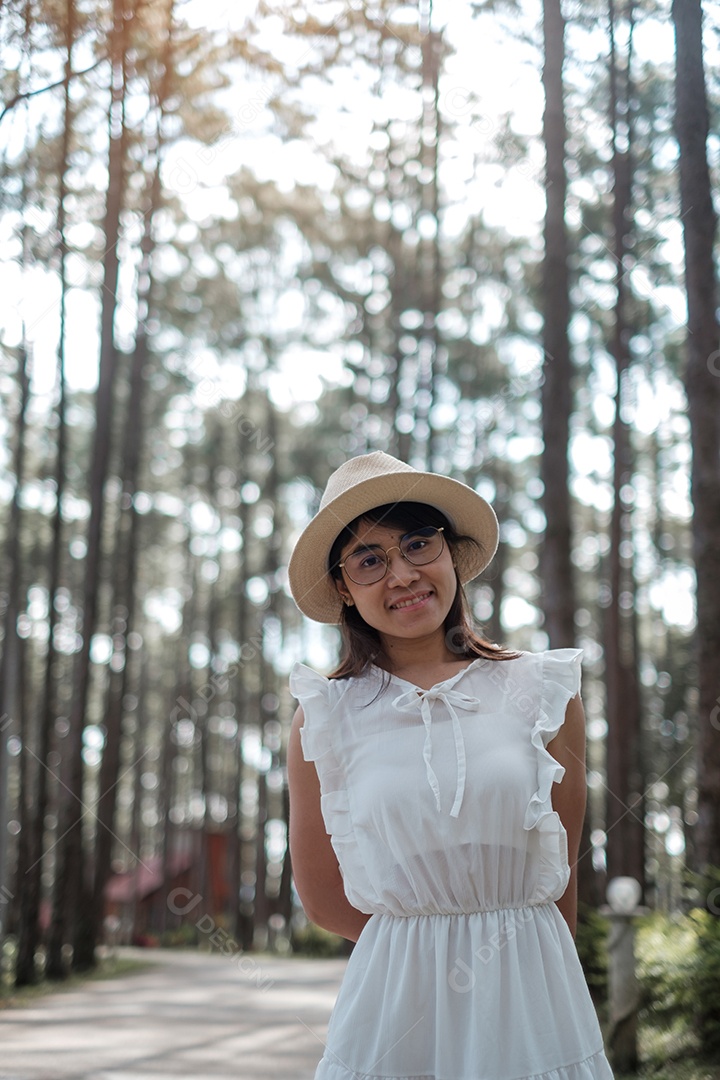 Mulher feliz de vestido branco e chapéu viajando na floresta de pinheiros.