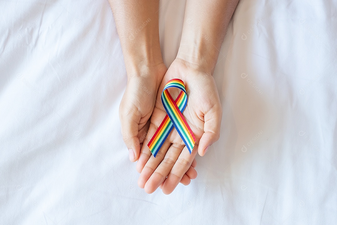 Mão mostrando a fita LGBTQ Arco-íris no fundo da cama branca.