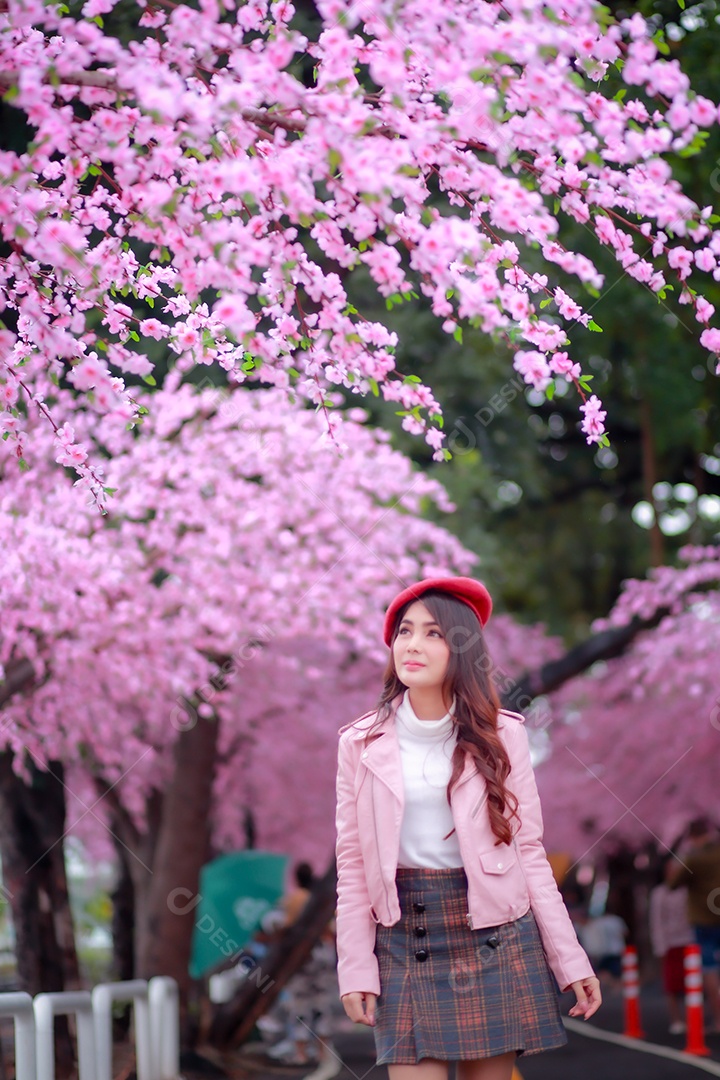 A hipster traveler woman strolling wears a red hat with a beautiful blossoming sakura cherry blossom tree in pink color in the park on a spring day.