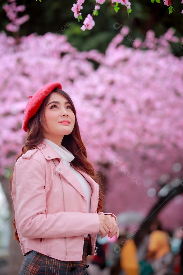 A hipster traveler woman strolling wears a red hat and a plain leather dress with a beautiful blossoming sakura cherry blossom tree in pink color in the park on a spring day.