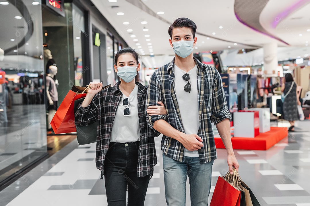 Jovem casal com máscara de proteção segurando várias sacolas de papel andando no corredor do grande shopping center