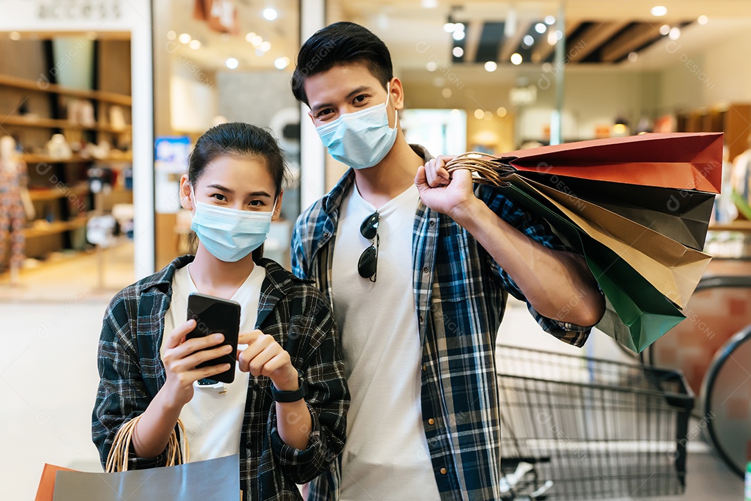 Jovem casal com máscara de proteção segurando várias sacolas de papel andando no corredor do grande shopping center