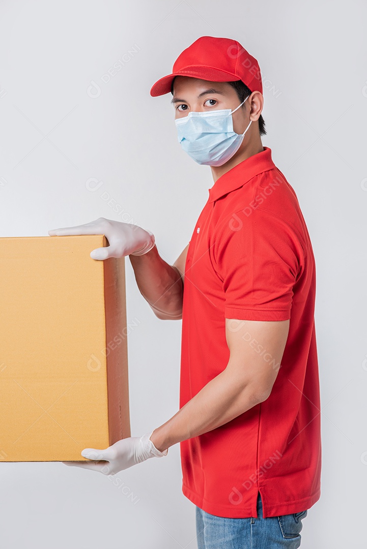 Imagem de um jovem entregador consciente de boné vermelho t-shirt em branco uniforme luvas de máscara facial em pé com caixa de papelão